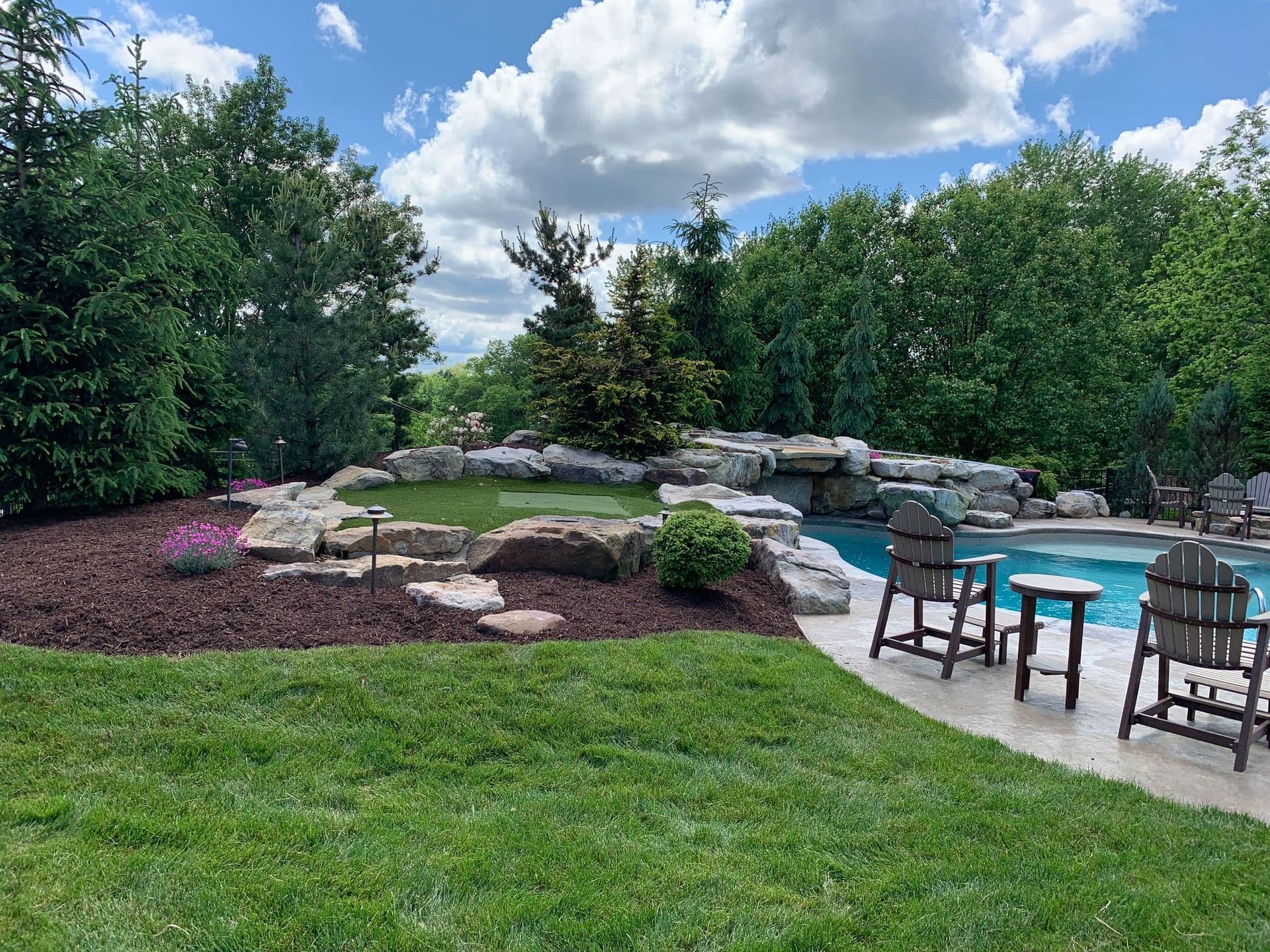 Beautiful landscaped backyard with a pool, rock formations, and seating area under a blue sky.